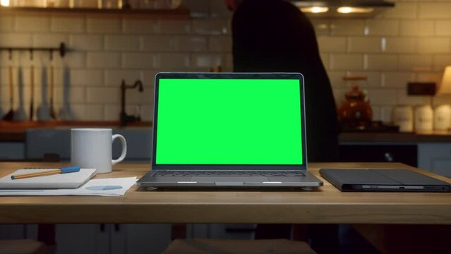 Laptop computer with mock up green screen chromakey display standing on the kitchen table. Anonymous person is preparing something in the background in kitchen at night.