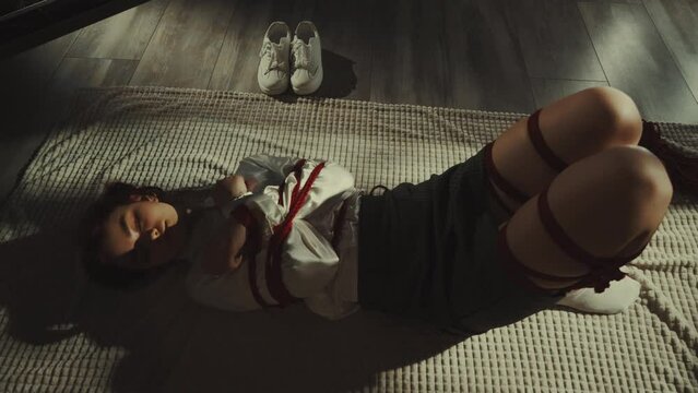 Zoom out: woman bound with red shibari ropes, lying on white blanket in softly lit room, traditional Japanese rope art form, aesthetics and calm. White sneakers placed nearby, intimate scene