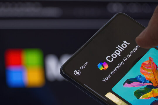 Copilot icon visible on mobile phone screen, Microsoft logo in the background. Copilot is a chatbot and artificial intelligence tool based on ChatGPT and developed by MS.