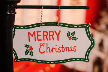 a sign advertising merry christmas hanging on a pole against a red christmas tree