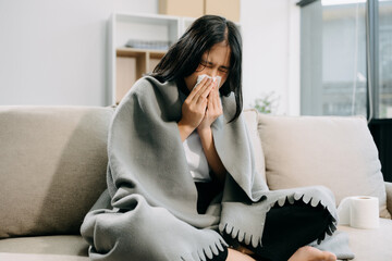 Young Asian woman suffering from flu symptoms, covered with a blanket on a sofa. Concept of...