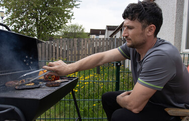 A young white man is barbecuing in the garden. BBQ. Meat and vegetables on the grill. Pepper, eggplant and meat patty for a burger on fire. A man turns meat over a fire with tongs