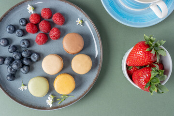 Macarons in a plate on a green background. Berries and pastries close-up with copy space. Strawberries, raspberries and blueberries are decorated with white flowers. French breakfast. Tea party