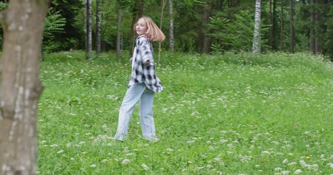 Excited teenage girl wearing checkered pattern casuals dancing amidst flowering plants. Happy female walking on grassy landscape in forest. She is spending leisure time during summer weekend.