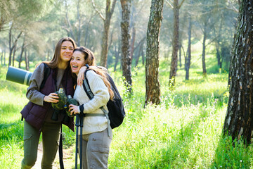 Two women are smiling and hugging each other while holding water canteen. They are doing trekking...