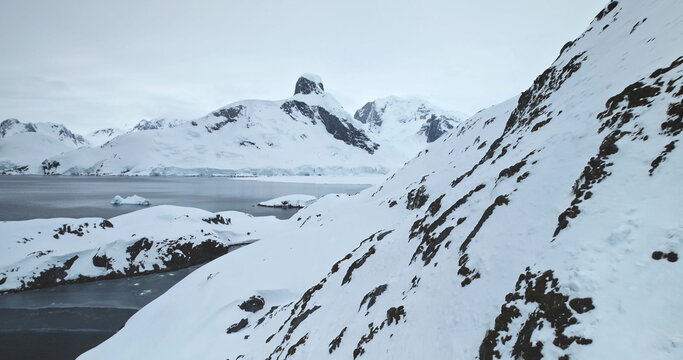 Fly over mountain snow covered rock in Antarctica. Arctic slope hill over cold polar ocean, mountain range peak in background. Harsh extreme low temperature conditions. South Pole explore and travel