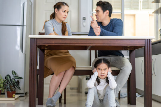 Family conflict concept, young couple arguing at the kitchen table while their little daughter sits on the floor covering her ears, distressed, mother and father are engaged in a heated discussion