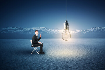 A man in a suit sitting and working on a laptop outdoors with a lightbulb overhead, snowy mountains...