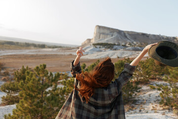 empowered woman wearing hat raises arms in front of majestic mountain range