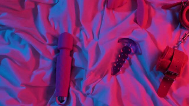 set of woman sex toys for masturbation and orgasm on sheet on bed. Female vibrator, vacuum stimulator, butt plug and BDSM handcuffs in neon lighting