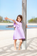 Happy little blonde girl in lilac summer dress playing and has fun on a white sandy beach near the...