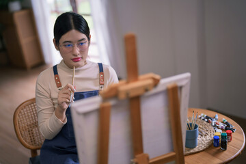 Young asian female artist hold paintbrush while looking on canvas to thinking idea painting artwork
