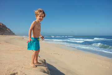 Cute boy on sandy beach smile and gaze at waves under blue sky