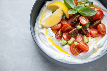 Bowl of greek feta dip topped with fresh tomatoes and cucumber, horizontal shot on a grey granite surface, middle closeup