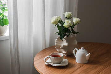 A cup of tea, a bouquet of white roses in a ceramic vase on a round wooden table in the living room. An aesthetic lifestyle