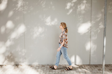 Side view of a young woman walking outdoors in casual attire on a sunny day, casting shadows on the wall.