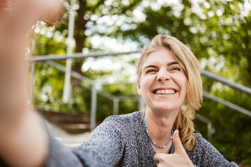 A cheerful woman smiling brightly and giving a thumbs up gesture while outdoors in a sunny park...