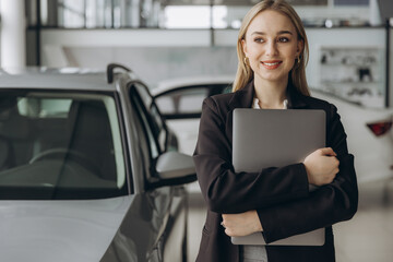 Pensive stylish businesswoman manager in suit holding laptop computer, standing near car at car dealership