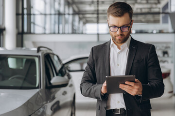 Bearded friendly car seller in glasses and suit standing in car salon and holding tablet