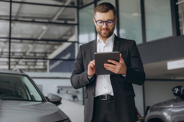 Bearded friendly car seller in glasses and suit standing in car salon and holding tablet