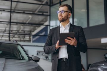 Bearded friendly car seller in glasses and suit standing in car salon and holding tablet