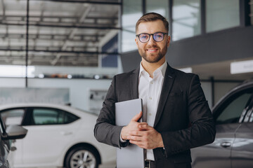 Modern bearded man in glasses and suit vehicle sales consultant using laptop inside car dealership