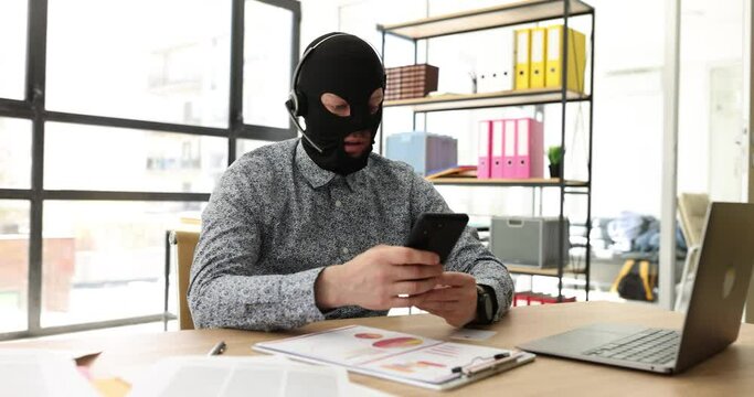 Male thief in a balaclava in headphones with microphone with phone in office. Cybercrime and phone scammers