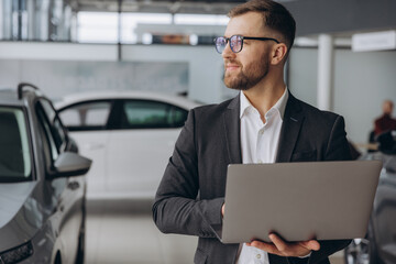 Modern bearded man in glasses and suit vehicle sales consultant using laptop inside car dealership