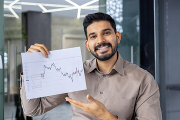 A man with a smile presents financial data on a chart in a modern office, symbolizing business...