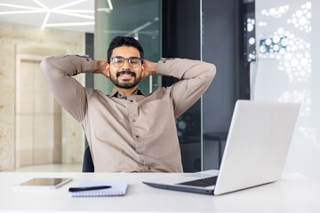 Fototapeta premium A man in an office setting is seen relaxing at his desk, surrounded by modern technology and office supplies. He appears calm and confident in his professional workspace