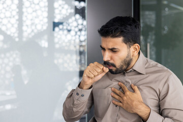 The image depicts a man in discomfort while holding his chest and coughing in a workplace setting....