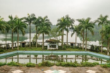 Deserted resort with palm trees, lake, and distant structures. Pool filled with murky water and garbage. Opposite shore has hotel-like buildings among trees. Abandoned Waterpark in Hue, Vietnam.