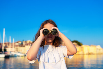 Funny little girl looking through vintage binoculars on sunny summer day. Rovinj town in background. Travel and adventure concept.