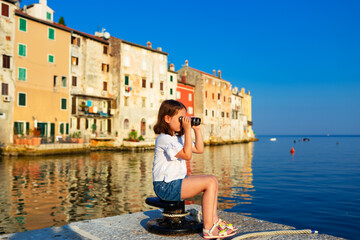 Funny little girl looking through vintage binoculars on sunny summer day. Rovinj town in background. Travel and adventure concept.