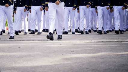 A group of cadets dressed in formal uniforms, marching in synchronized formation on a paved road during a military parade or drill.