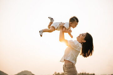 In a carefree summer moment, mom holds her cute toddler son up high, throwing him up into sky. playful child enjoys freedom of flying, while cheerful mother smiles and enjoy moment of family happiness