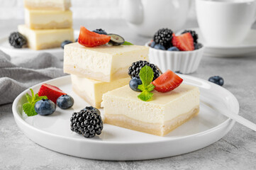 Vanilla cheesecake cut into squares with fresh berries on a white plate on a gray stone background