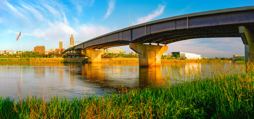 Omaha City Skyline, I-480 Missouri River Bridge, and gentle water flow at warm glowing sunrise: A tranquil summer landscape from Council Bluffs Riverfront in Iowa 