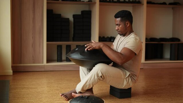 Black male in trance practicing native music traditional drumming instrument playing with hands sitting cross-legged on floor. Serene African man drumming rhythm on steel tongue drum by hands, slowmo.