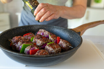 Woman preparing delicious minced meat skewers in a frying pan and season with pepper
