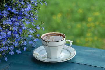 Cup of coffee on a blue table in balcony near by lobelia in a pot on blurred background of green...