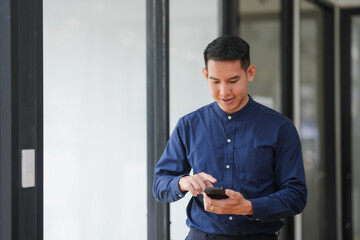 Businessman in a blue shirt using a smartphone in a modern office hallway, focusing on work-related tasks.