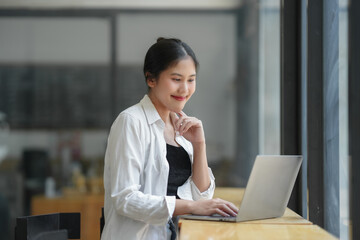 Young professional woman working on laptop with a smile, wearing a casual white shirt, seated near...