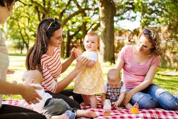 Mothers and babies enjoying group picnic outdoor in park, sitting on picnic blanket and preparing food and drinks.