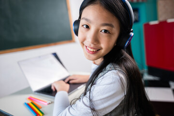 A young girl is sitting at a desk with a laptop and a book. She is smiling and wearing headphones