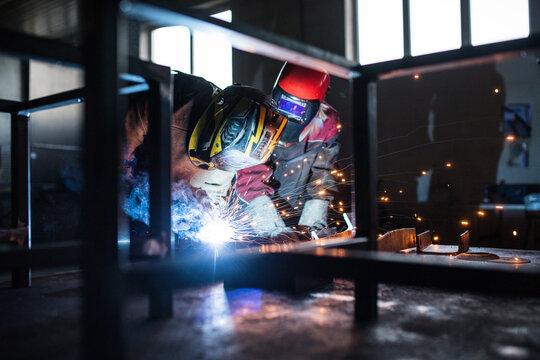 Blonde woman works as a welder in workshop, operating welding machine, wearing protective clothing and a welding mask. Two welders helpign each other.