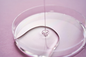 Serum or cosmetic oil flows into a transparent bowl on a purple background.