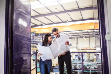 Two project managers standing in modern industrial factory. Manufacturing facility with robotics, robotic arms and automation. Storing products and materials in warehouse.