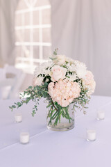 A simple floral arrangement in a vase, surrounded by flowers on a white tablecloth table in a tented wedding reception. 