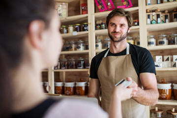 Handsome shop assistant serving customer in package-free store using reusable containers. Zero waste shops offering package-free bulk goods and sustainable alternatives.